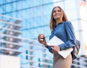 blonde young woman smiling portrait holding laptop coffee wearing blue gentle shirt modern building ()